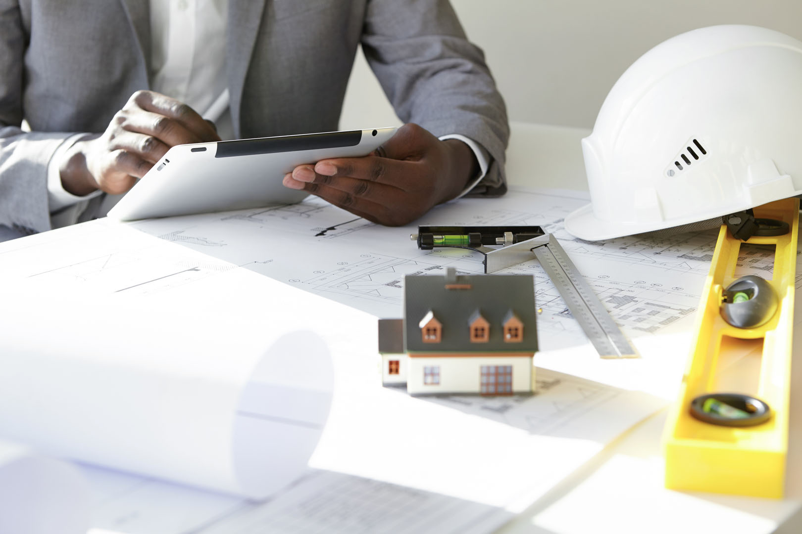Cropped shot of dark skinned contractor holding touch pad, entering data while working on new housing project, sitting at desk with drawings, scale model house, blueprint rolls, ruler and helmets